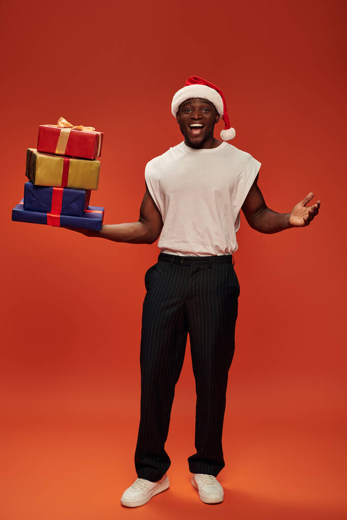 возбужденный африканский американец в шапке Санты держит подарочные коробки и показывает вау жест на красном фоне - Фото, изображение