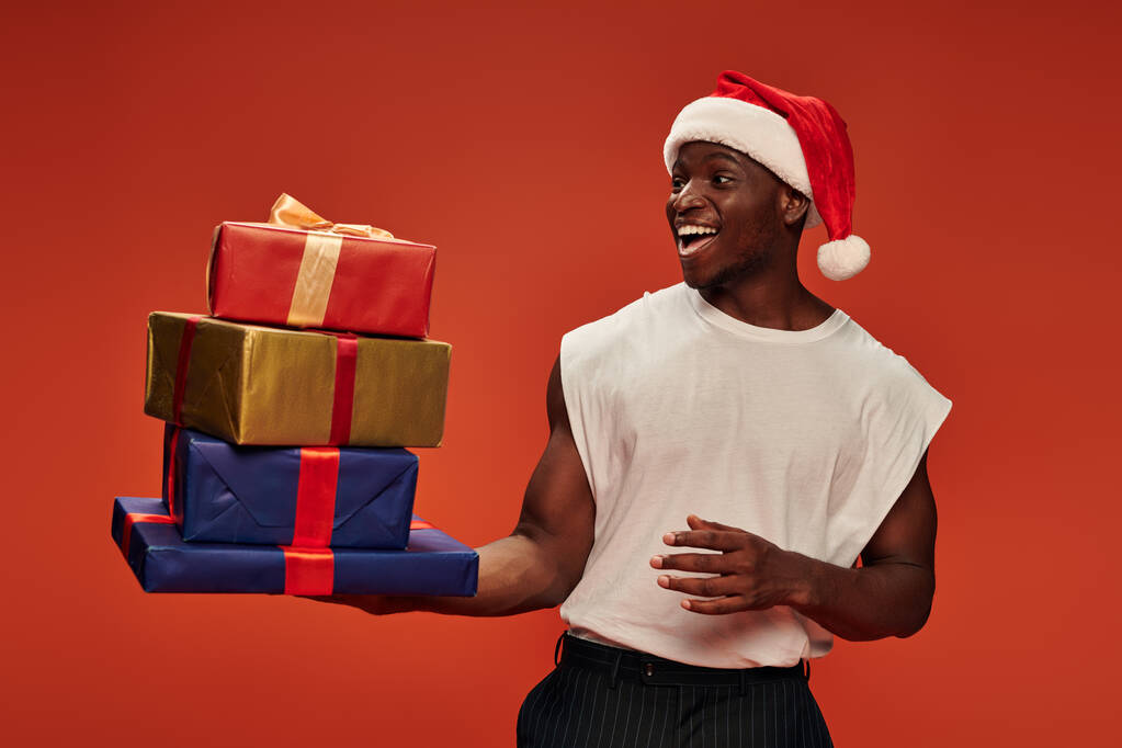 erstaunt und überglücklich afrikanisch-amerikanischer Mann mit Weihnachtsmütze schaut auf bunte Geschenkboxen auf rot - Foto, Bild