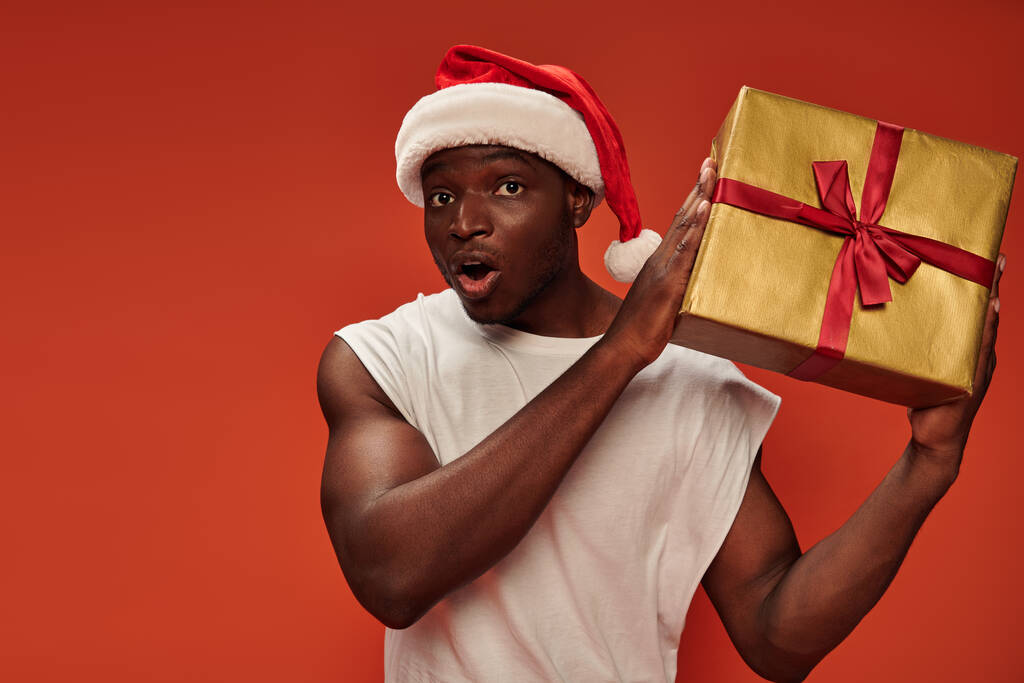 zaskoczony Afroamerykanin człowiek w Santa kapelusz trzyma pudełko prezentów i patrząc na aparat fotograficzny na czerwonym tle - Zdjęcie, obraz