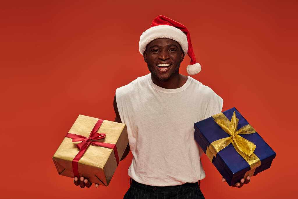 クリスマスの帽子とホワイトタンクトップで喜びのアフリカ系アメリカ人男性 赤でお祝いのプレゼントをポーズ - 写真・画像