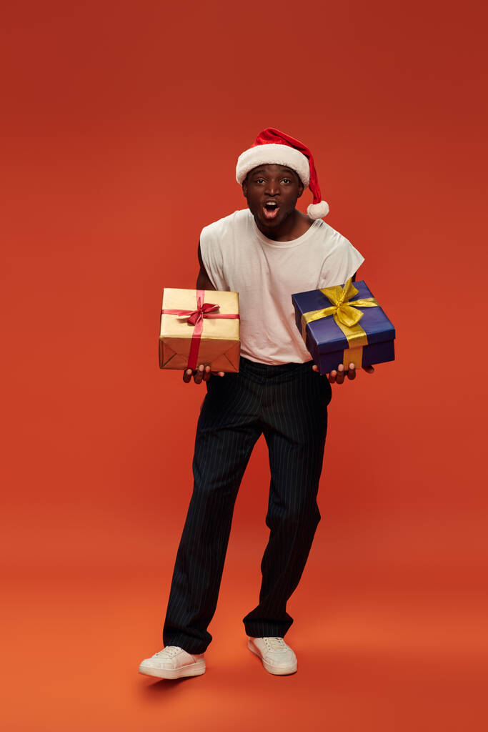 схвильований афроамериканський чоловік у повсякденному вбранні та капелюсі Санти, що тримає різдвяні подарунки на червоному - Фото, зображення