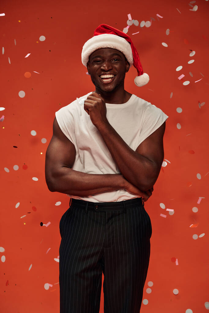 vreugdevolle Afrikaanse Amerikaanse man in kerst hoed kijken naar camera op rode achtergrond met vallende confetti - Foto, afbeelding
