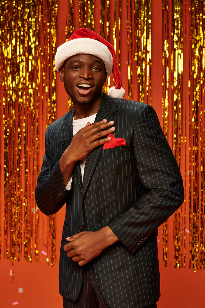 кокетливый африканский американец в элегантном костюме и кепке Санты, смотрящий в камеру рядом с золотой мишурой - Фото, изображение