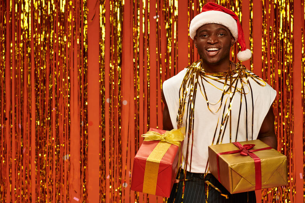 gelukkig Afrikaans amerikaanse man in santa hoed met geschenkdozen in de buurt van gouden tinsel op oranje achtergrond - Foto, afbeelding