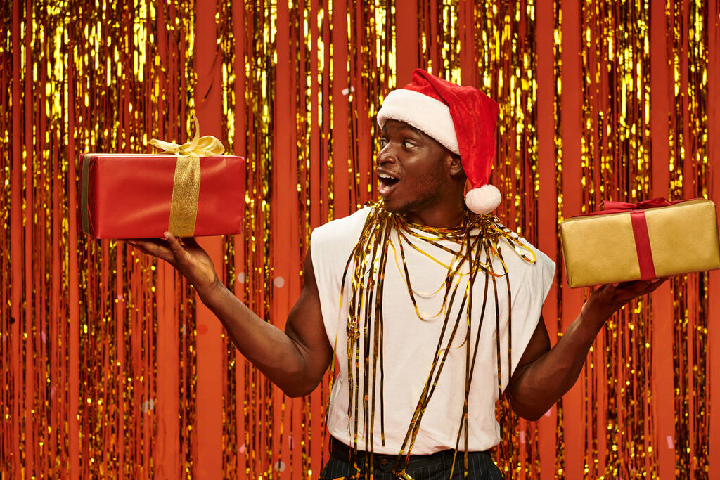 homme afro-américain étonné en chapeau de Père Noël avec des cadeaux de Noël près de tinsel brillant sur fond rouge - Photo, image