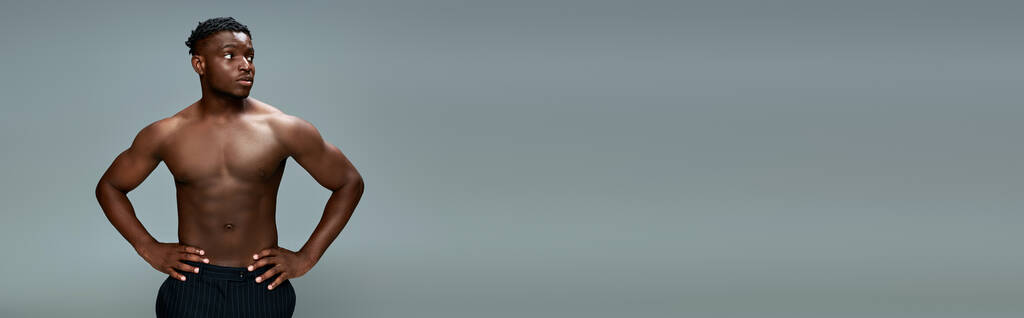 Üstsüz ve kaslı Afro-Amerikan erkeği kalçalarını tutuyor ve gri pankarta bakıyor. - Fotoğraf, Görsel