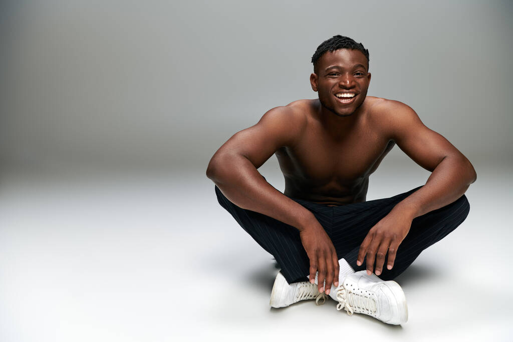 fröhlicher afrikanisch-amerikanischer Typ mit starkem, hemdlosen Körper, der auf grauen Ganzkörperanzügen sitzt und lächelt - Foto, Bild