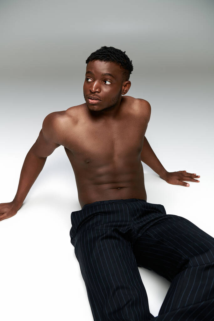 若いシャツレスのアフリカ系アメリカ人男性 筋肉トルソ座って灰色の背景を見つめている - 写真・画像
