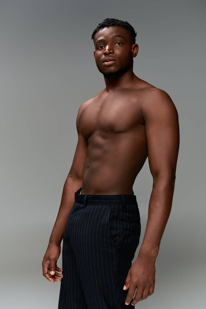 shirtloze Afrikaanse Amerikaanse fitness model in zwarte broek staan en kijken naar camera op grijs - Foto, afbeelding