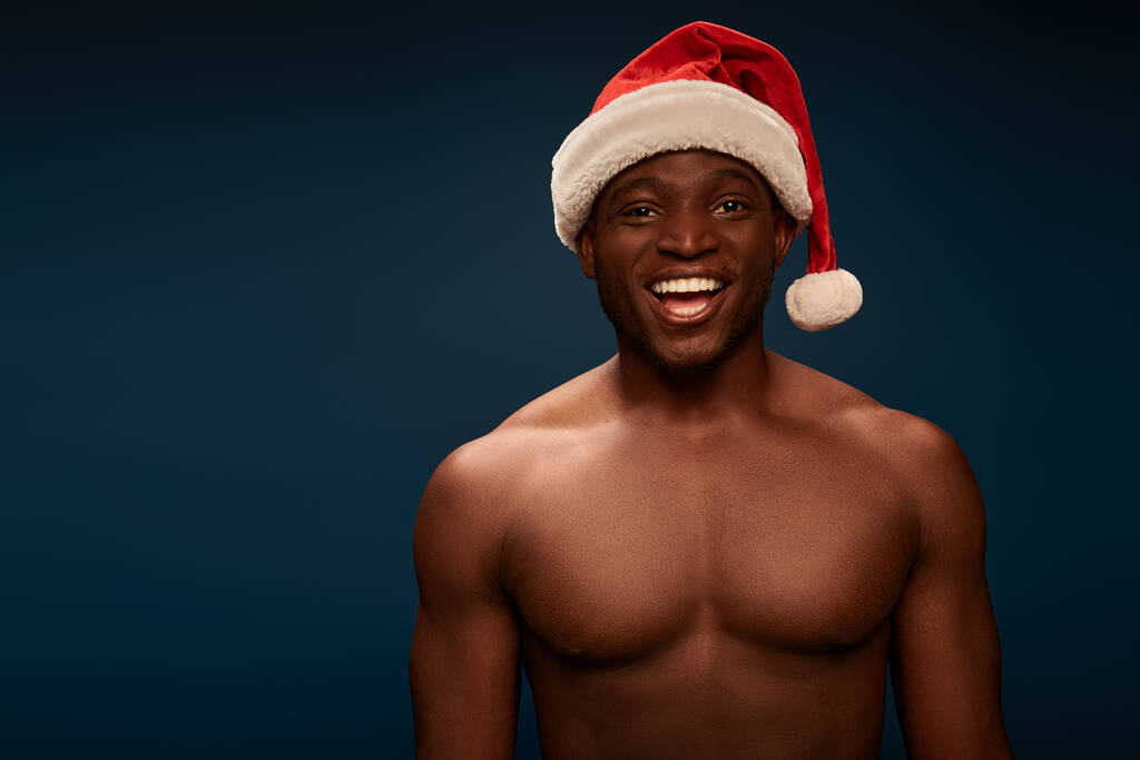 gelukkig en shirtless Afrikaans amerikaan guy in santa hoed glimlachen op camera op marine blauw achtergrond - Foto, afbeelding