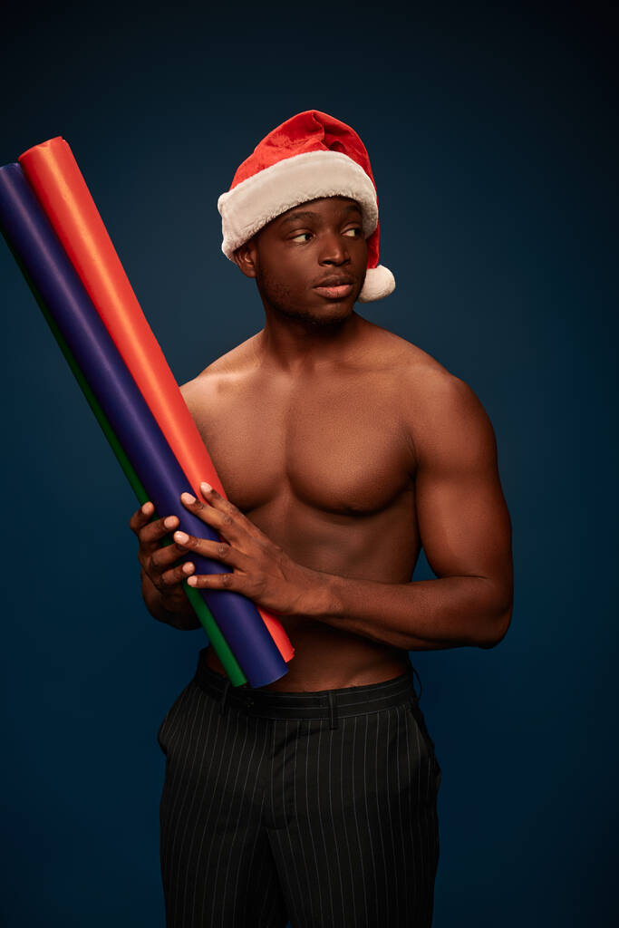 hemdloser afrikanisch-amerikanischer Typ mit Weihnachtsmütze, der bunte Papierrollen auf dunkelblauem Hintergrund hält - Foto, Bild
