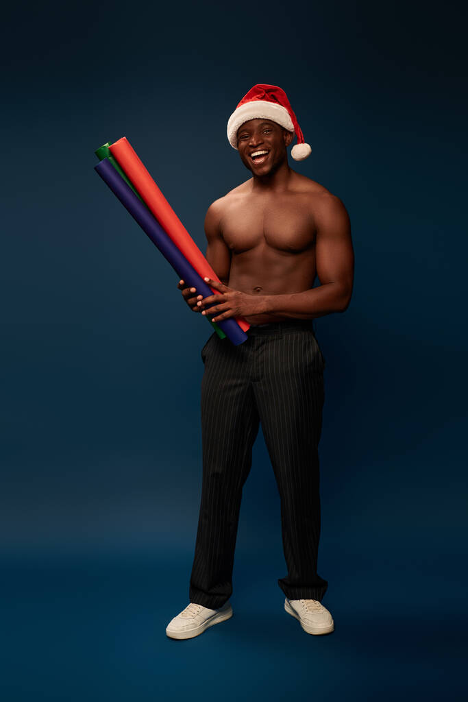 alegre musculoso afroamericano hombre en sombrero de santa con fondos de pantalla de colores sobre fondo azul marino - Foto, Imagen