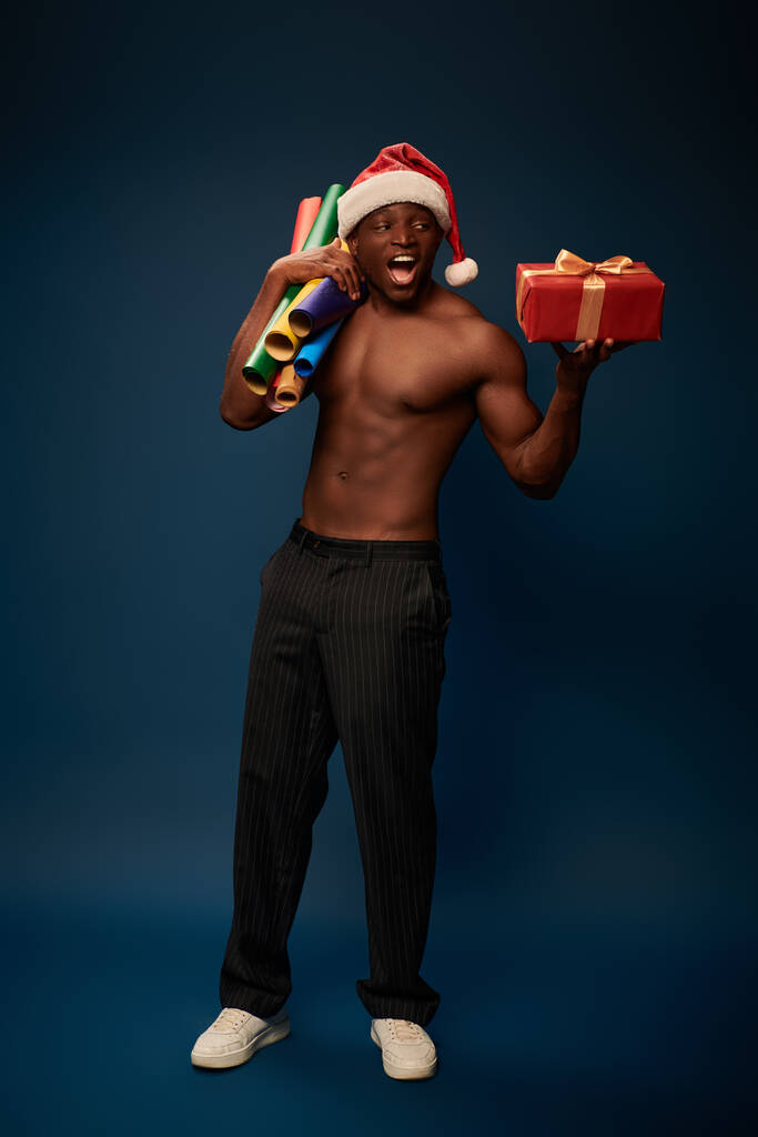 homme afro-américain excité et sportif en chapeau de Père Noël avec boîte cadeau et rouleaux de papier sur bleu foncé - Photo, image