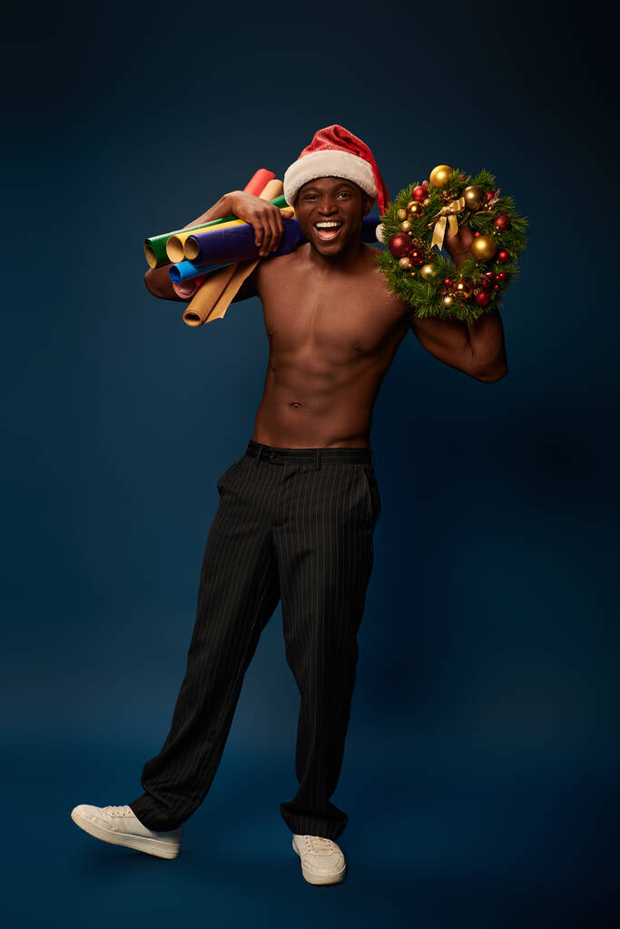 sportowy Afrykański Amerykanin w kapeluszu Mikołaja z tapetami i świątecznym wieńcem uśmiechnięty na granatowym tle - Zdjęcie, obraz