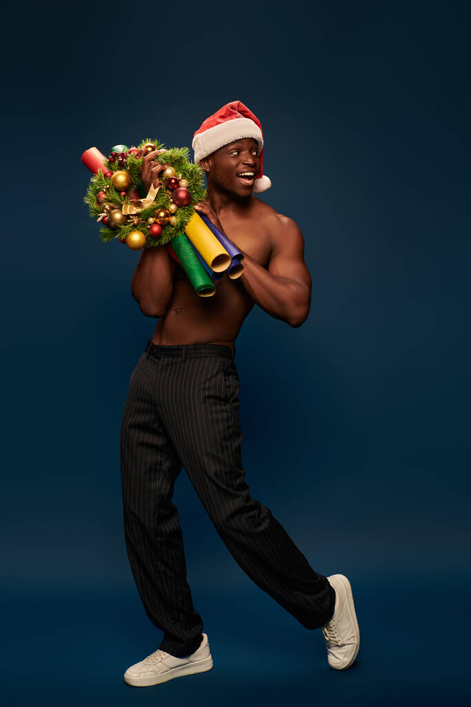 joyeux homme afro-américain torse nu en santa chapeau de marche avec décor de Noël sur bleu marine - Photo, image