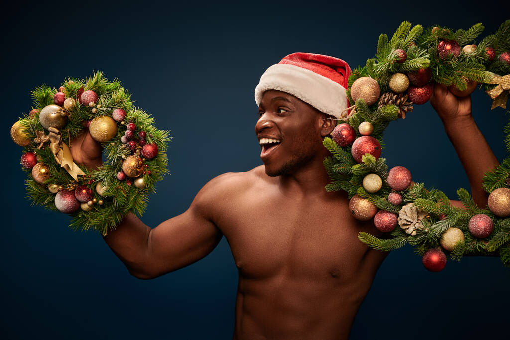 excited shirtless african american man in santa hap standing with christmas wreaths on dark blue - Photo, Image