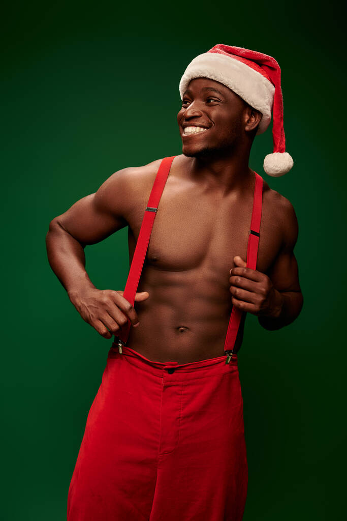 aufgeregter Afroamerikaner mit Weihnachtsmütze und roter Hose, der auf grünem Hintergrund wegschaut - Foto, Bild