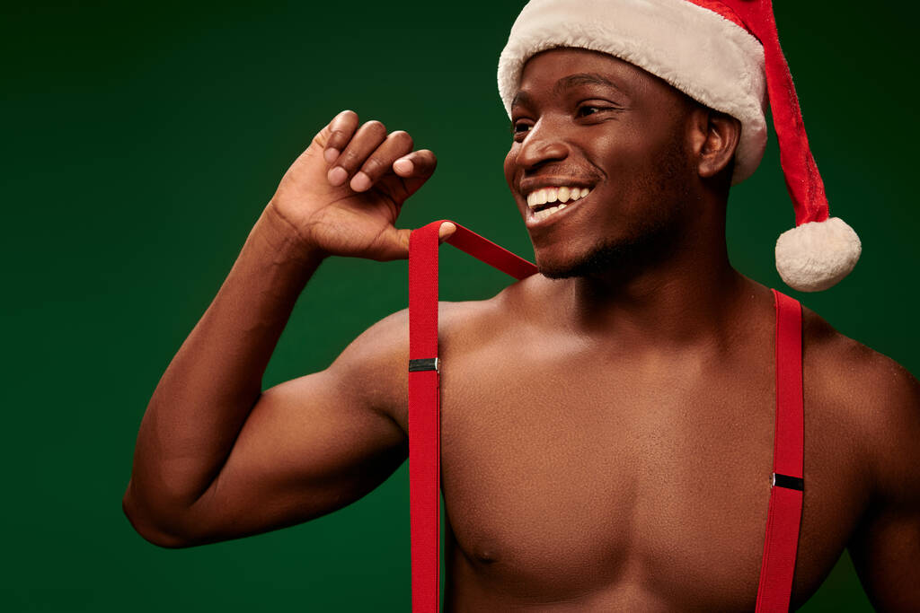 спортивний афроамериканський чоловік у капелюсі Санти і підтяжки посміхаючись і дивлячись на зелений - Фото, зображення