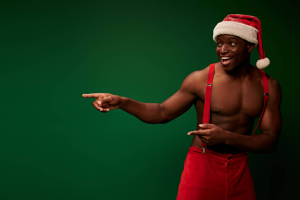 hombre afroamericano sin camisa en sombrero de santa y pantalones rojos sonriendo y señalando hacia fuera en verde - Foto, Imagen