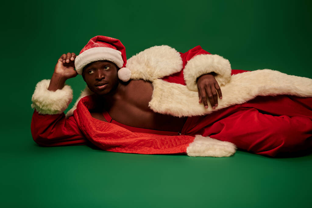 σέξι Αφροαμερικανός άνδρας με κοστούμι Άγιος Βασίλης σε γυμνό σώμα ξαπλωμένος και κοιτάζοντας μακριά στο πράσινο - Φωτογραφία, εικόνα