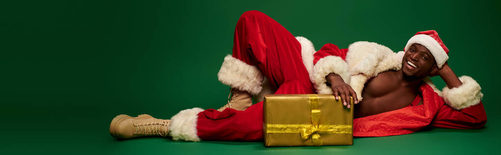 χαμογελαστός σέξι Αφροαμερικάνος με κοστούμι Άγιος Βασίλης ξαπλωμένος κοντά σε γυαλιστερό κουτί δώρου στο πράσινο, λάβαρο - Φωτογραφία, εικόνα