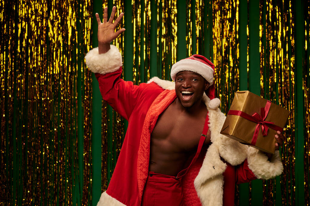 jolly afro-amerikai férfi karácsonyi jelmez fogja jelen és integető kéz fényes háttér - Fotó, kép