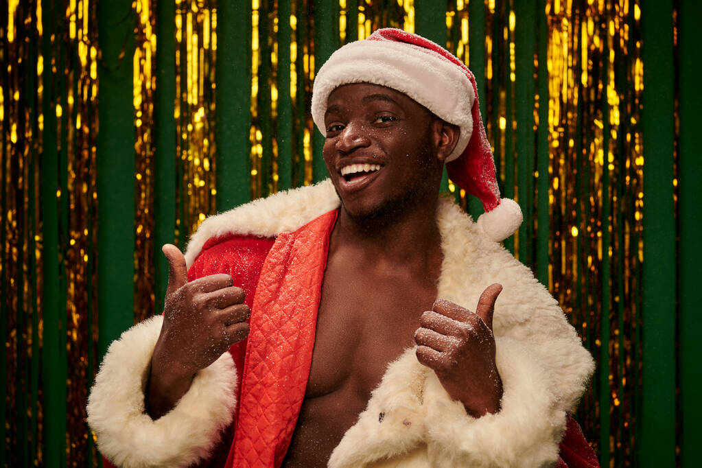 καυτό σκούρο δέρμα τύπος με κοστούμι Σάντα σε shirless σώμα δείχνει τους αντίχειρες επάνω στο φόντο των Χριστουγέννων - Φωτογραφία, εικόνα