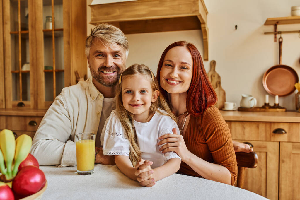 šťastní rodiče s roztomilou dcerou při pohledu na kameru v blízkosti čerstvého ovoce a pomerančové šťávy v kuchyni - Fotografie, Obrázek