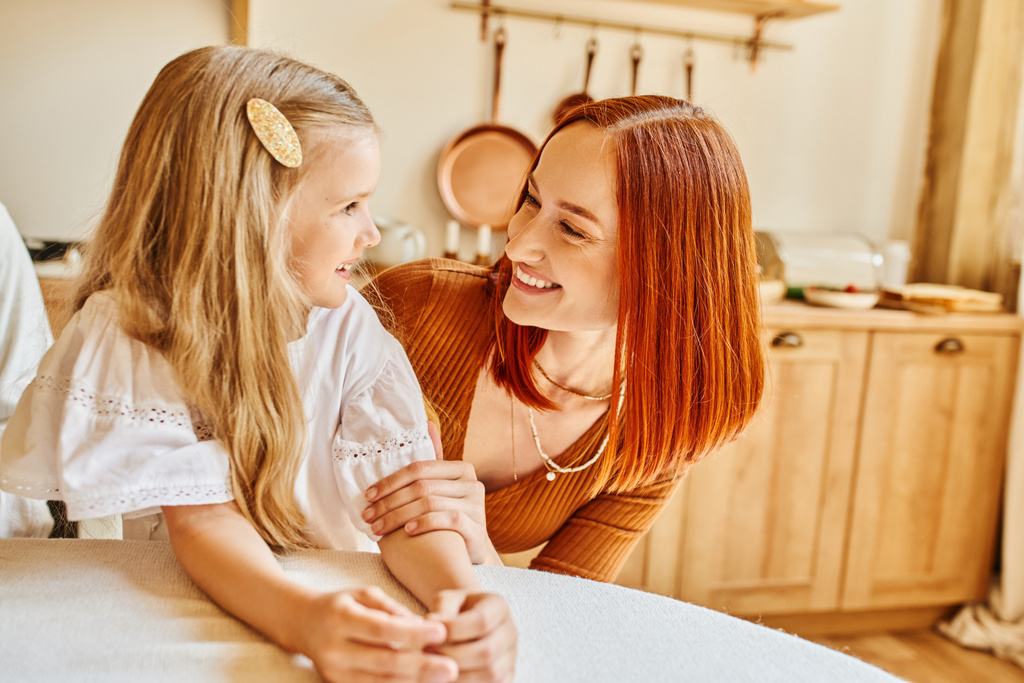 cheerful redhead woman and cute daughter looking at each other in cozy kitchen, love and care - Photo, Image