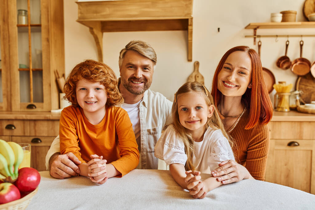 Lächelnde Eltern mit glücklichen Kindern, die in der gemütlichen Küche neben frischem Obst in die Kamera schauen, freudige Momente - Foto, Bild