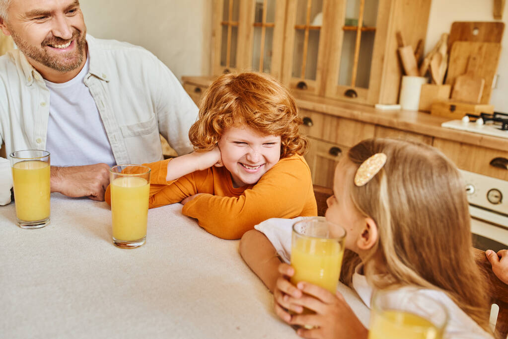 zorgeloze broer en zus glimlachend naar elkaar in de buurt van vers sinaasappelsap en ouders in de keuken thuis - Foto, afbeelding