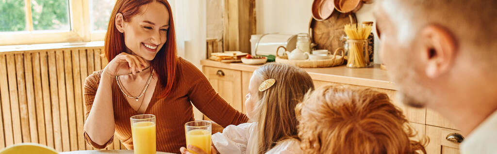 χαρούμενη γυναίκα κοιτάζοντας τα παιδιά κοντά σε φρέσκο χυμό πορτοκαλιού κατά τη διάρκεια του πρωινού στη ζεστή κουζίνα, πανό - Φωτογραφία, εικόνα
