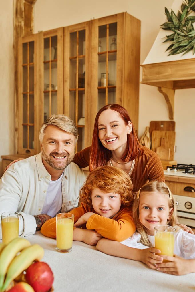 pleased couple with adorable kids looking at camera near fresh fruits and orange juice in kitchen - Photo, Image