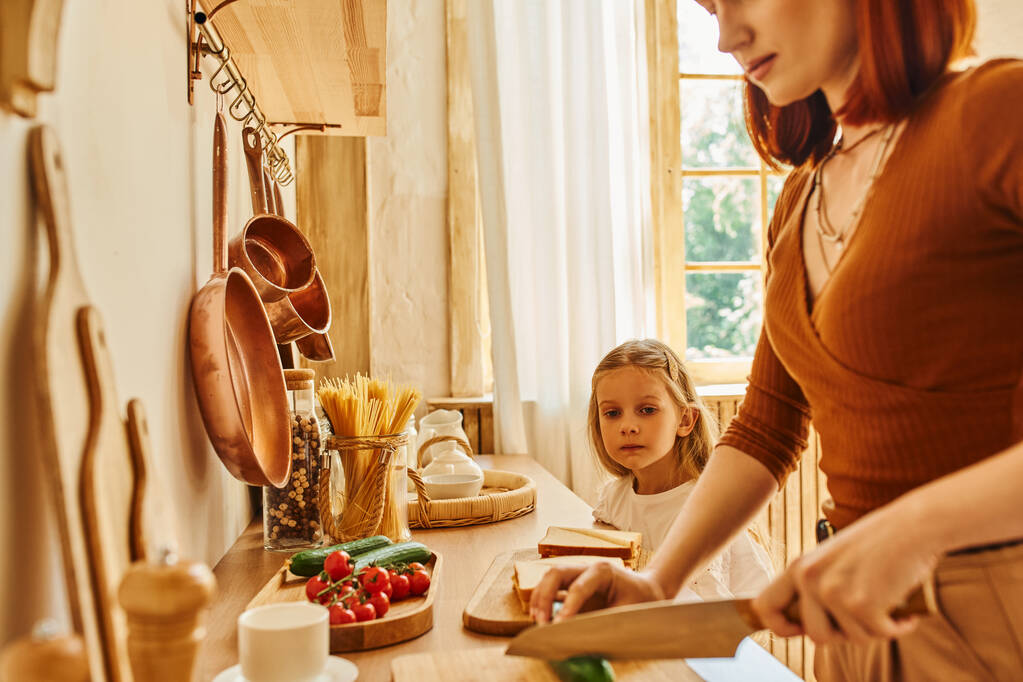 Lächelnde Mutter schneidet Gemüse bei der Frühstückszubereitung neben Tochter mit Sandwich auf der Küchentheke - Foto, Bild