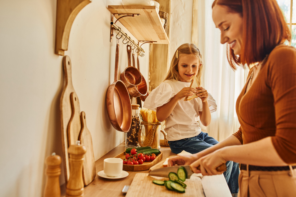 счастливая дочь сидит с бутербродом на кухне счетчик рядом улыбается мама готовит завтрак - Фото, изображение
