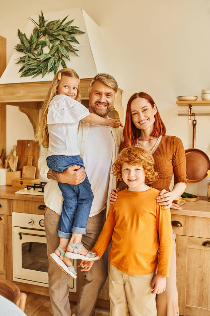 padres alegres con niños adorables mirando a la cámara en la acogedora cocina moderna, conexión emocional - Foto, imagen