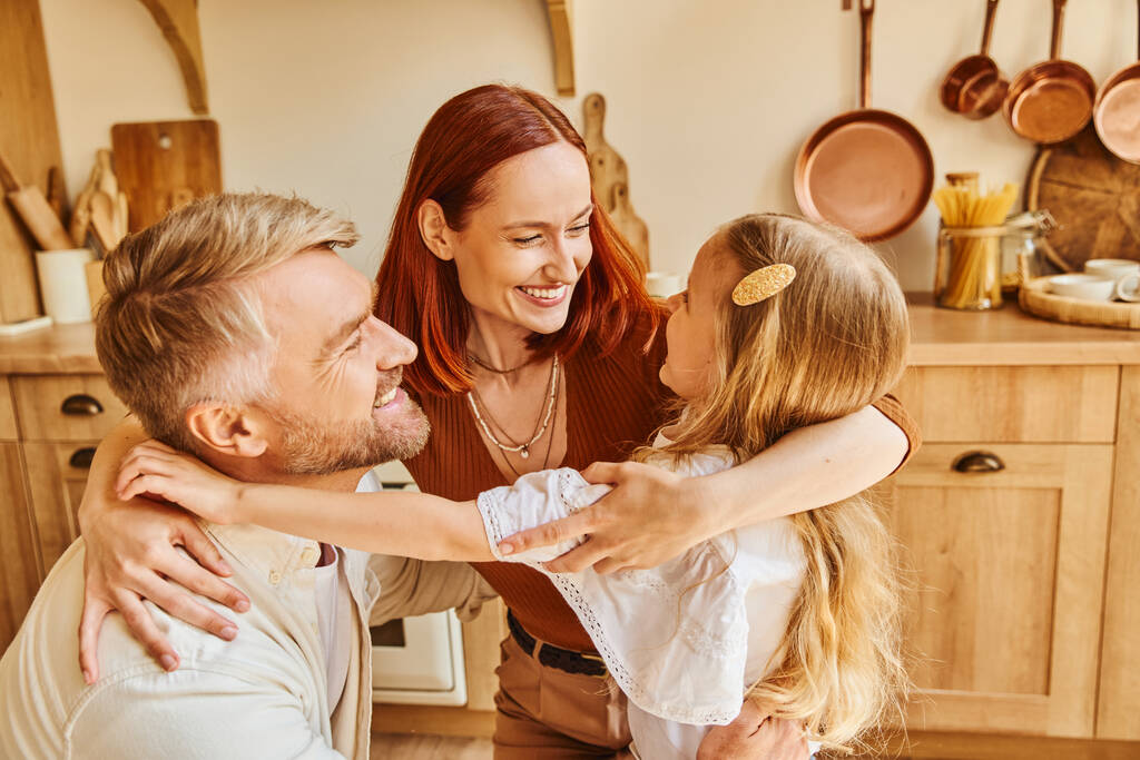 любящие родители с очаровательной дочерью обнимаются в уютной кухне дома, связывая семейные моменты - Фото, изображение
