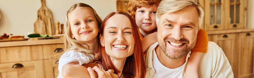 glimlachende ouders met gelukkige kinderen omarmen en kijken naar camera in gezellige keuken, horizontale banner - Foto, afbeelding