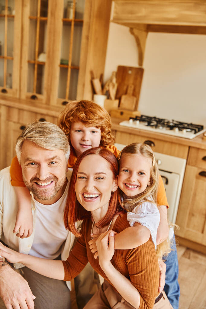 счастливые родители и дети, обнимающиеся и смотрящие в камеру на уютной кухне, заветные моменты - Фото, изображение