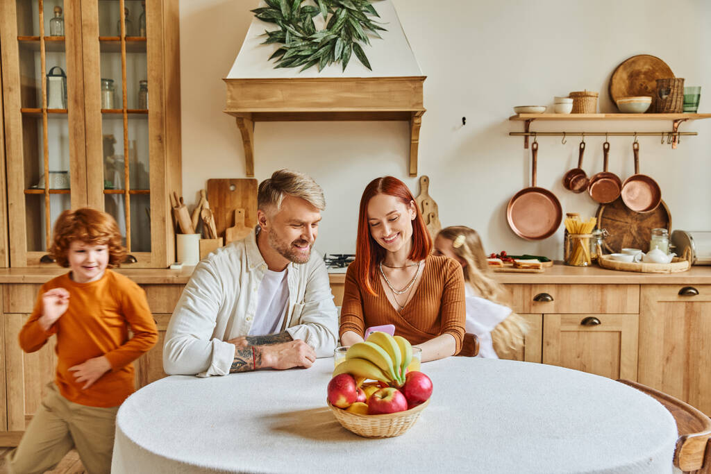 játékos gyerekek rohangálnak boldog szülők ül okostelefon közelében friss gyümölcsök hangulatos konyhában - Fotó, kép