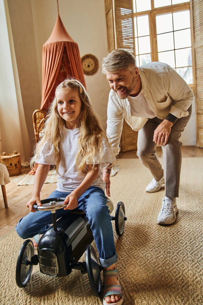 happy man assisting cheerful daughter riding toy car in modern living room at home, playing together - Photo, Image