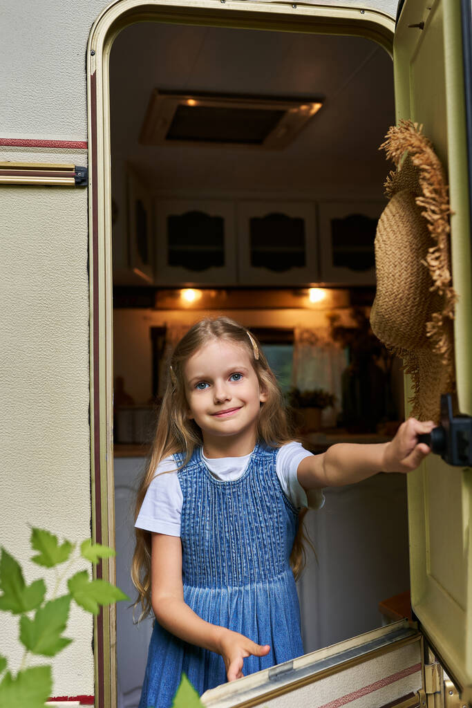 blondynka dziewczyna w dżinsowe sukienka otwarcie okna na przyczepie domu i patrząc na aparat fotograficzny, szczęśliwy dzieciństwo - Zdjęcie, obraz