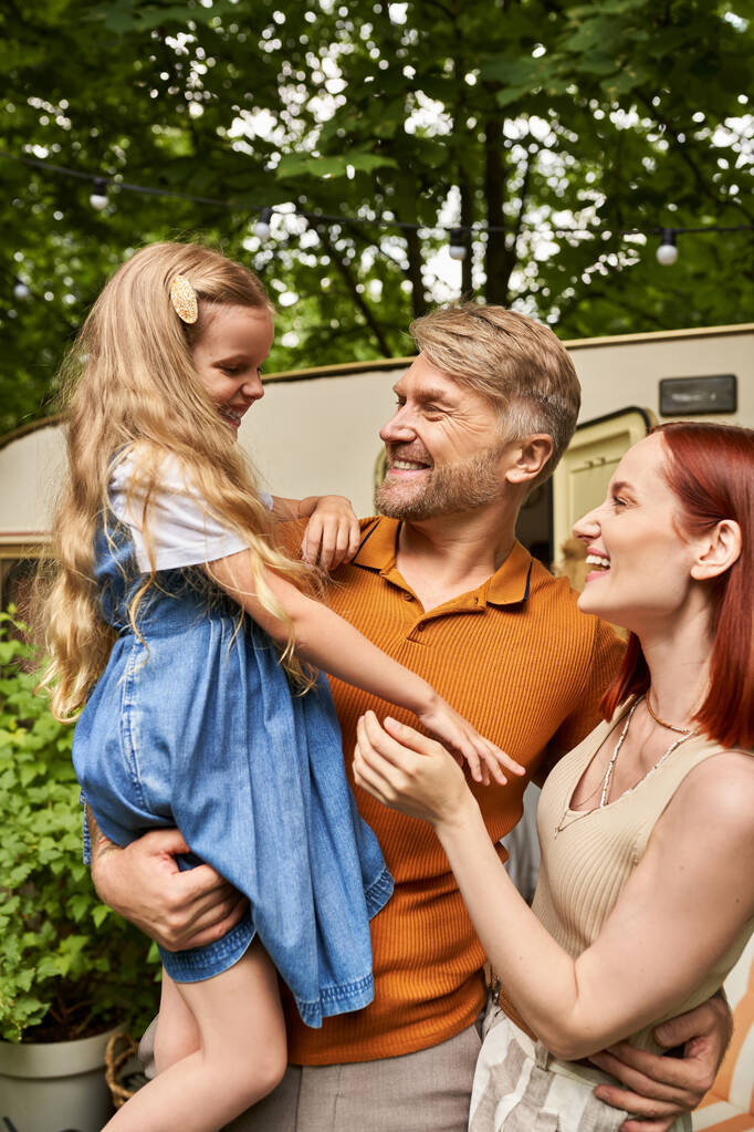 pai segurando alegre filha perto da esposa satisfeita e moderna casa reboque, lazer e aventura - Foto, Imagem