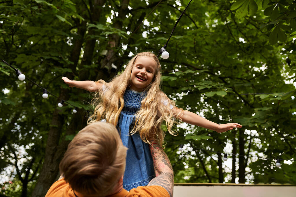 татуированный мужчина держит и играет с веселой дочерью в зеленом парке, счастливые семейные моменты - Фото, изображение