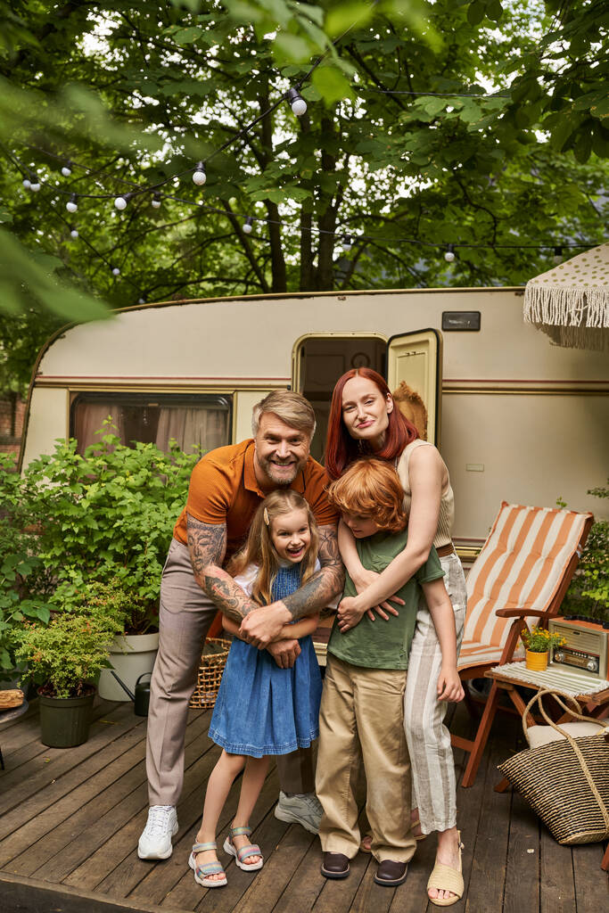 happy parents embracing cheerful children near modern home on wheels in trailer park, family leisure - Photo, Image