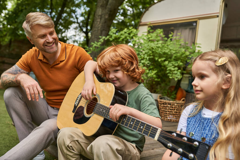 pelirroja tocando la guitarra cerca de sonreír padre y hermana al lado de casa remolque, actividades creativas - Foto, imagen
