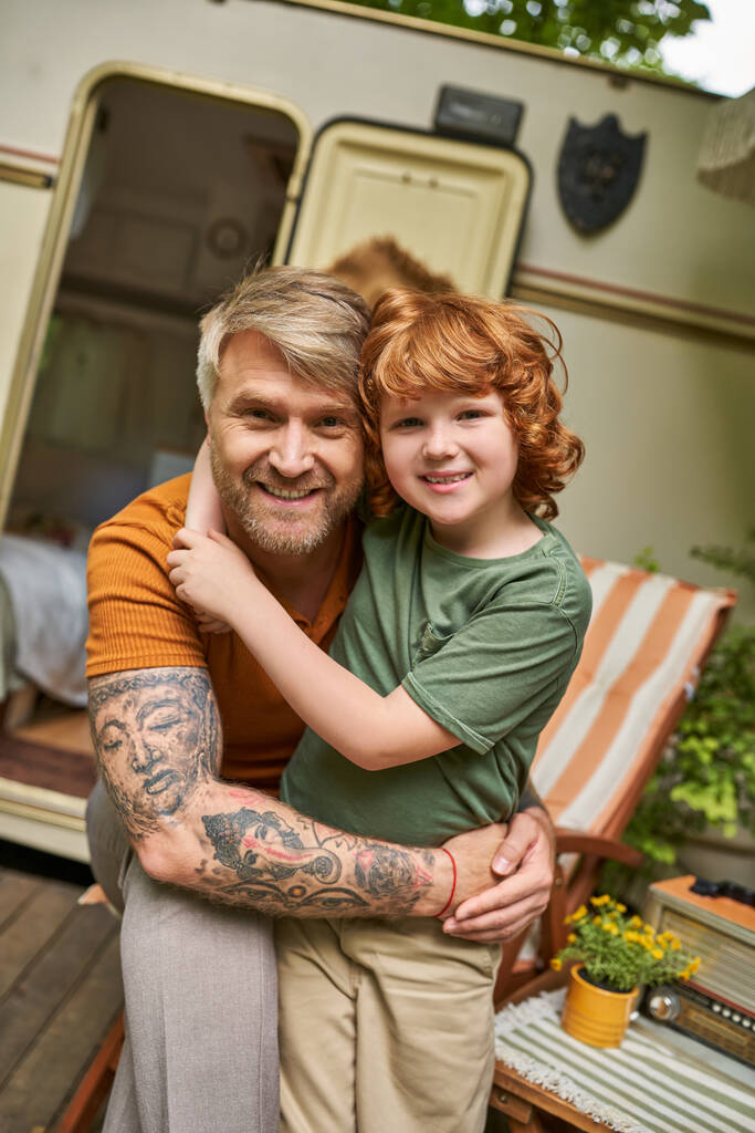 joyful tattooed man with adorable redhead son embracing and smiling at camera next to trailer home - Photo, Image