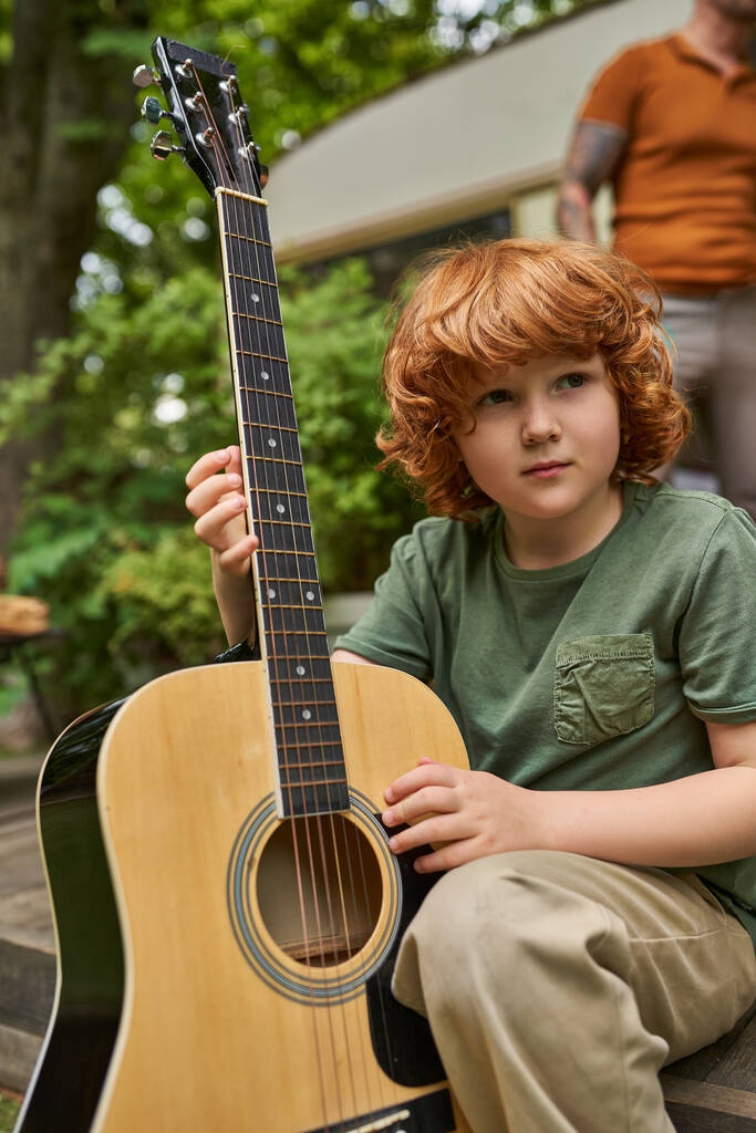 現代のトレーラーの家の近くに座っている間,アコースティックギターを離れて見ている夢のような赤毛の少年 - 写真・画像