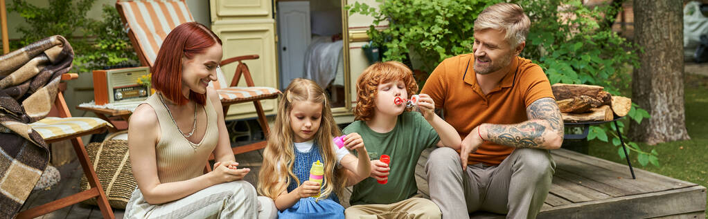 uśmiechnięci rodzice patrzący na dzieci dmuchające bańki mydlane w pobliżu przyczepy domowej, rozrywka i zabawa, baner - Zdjęcie, obraz