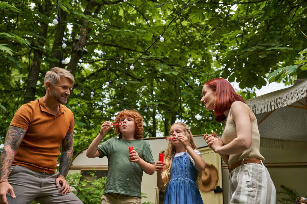 smiling parents looking at adorable children blowing soap bubbles near trailer home outdoors - Photo, Image